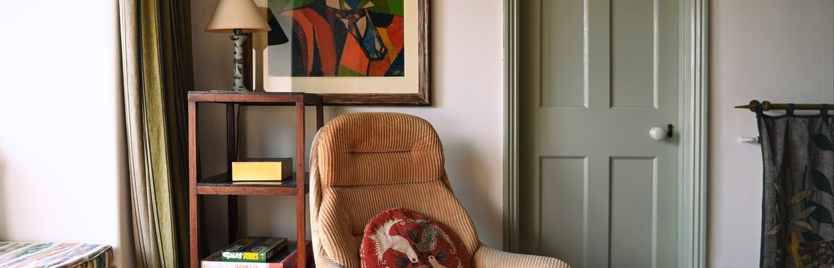 Living room scene with cosy armchair in the centre, bookshelf with table lamp, artwork and green door in the background