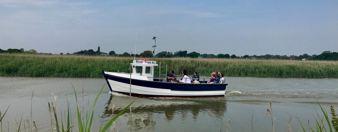 Small touring boat motoring down the river, with reeds and fields in the background.